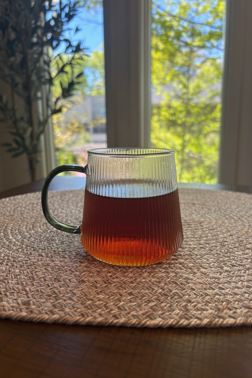 Glass mug with a dark liquid on a textured surface, with a window in the background showing greenery.